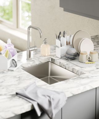 A sleek kitchen setup featuring a stainless steel undermount sink with a marble-effect countertop. The sink is complemented by a brushed chrome tap. Nearby, a dish rack holds plates, mugs, and cutlery, while a soap dispenser and sink caddy with cleaning brushes provide functionality.