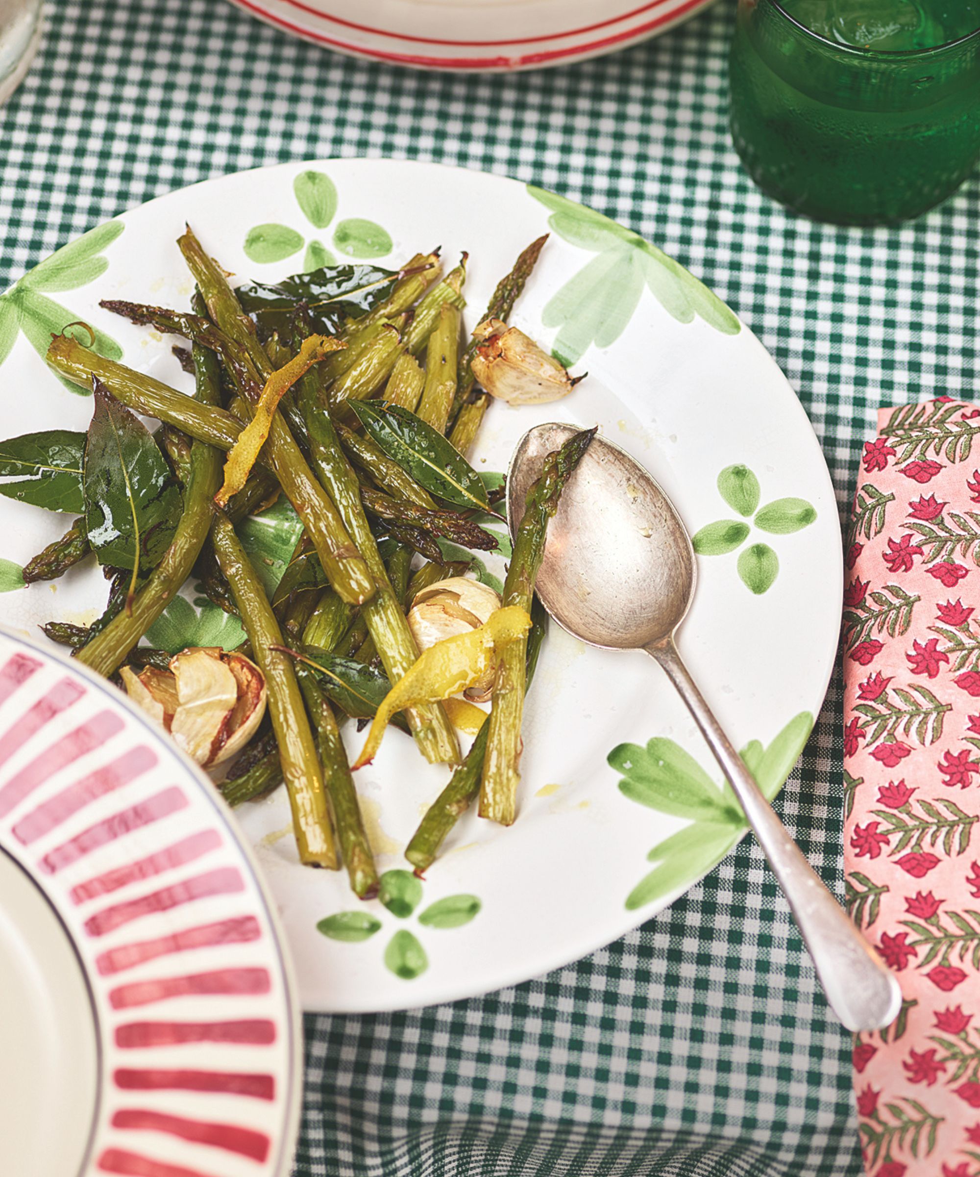 Asparagus with bay leaves and whole crushed garlic cloves on a white plate with green patterns and a table with a green and white gingham tablecloth, a pink floral napkin and a pink and whire striped plate