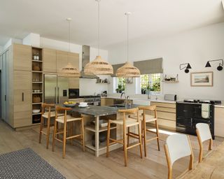 A large kitchen with wooden flooring, cabinets and bar stools around a central island below rattan lights