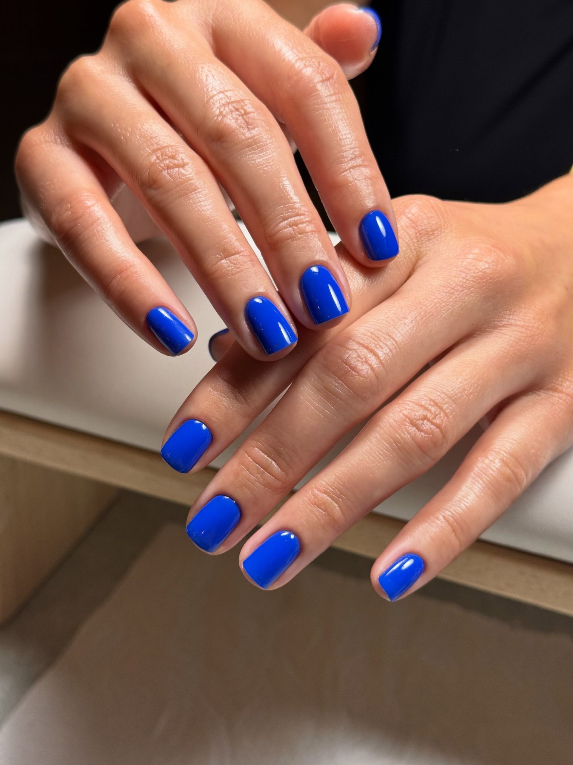 Two hands with short, glossy cobalt blue nails rest against a white surface.
