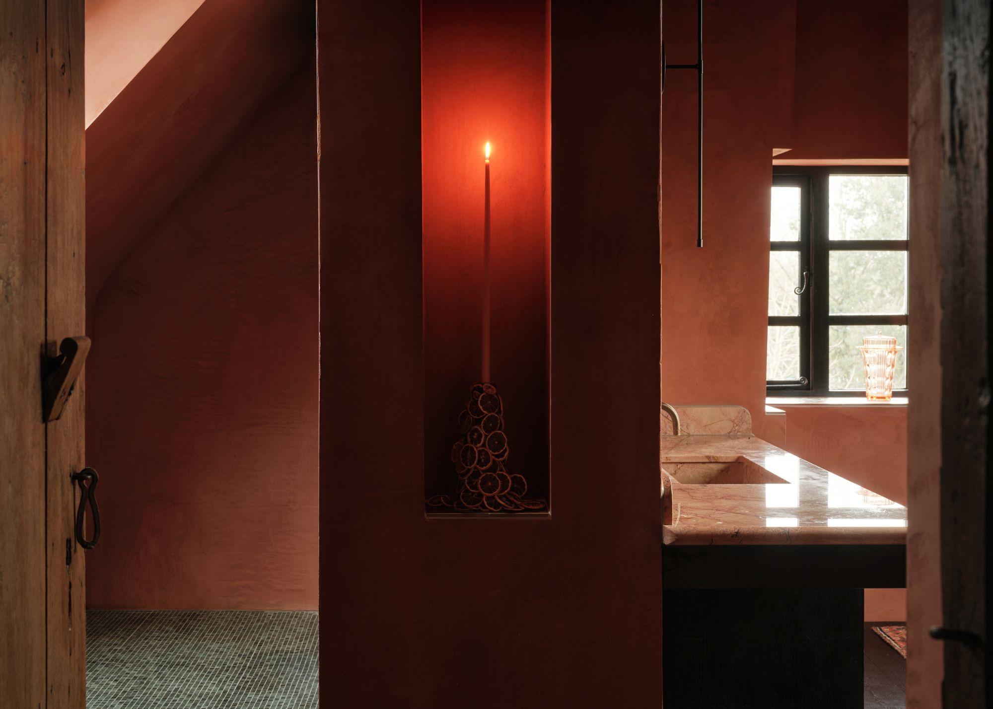 A red bathroom with a wall that holds a candle in the middle. There is a slight view of the marbled bathroom sink and the window within the bathroom.
