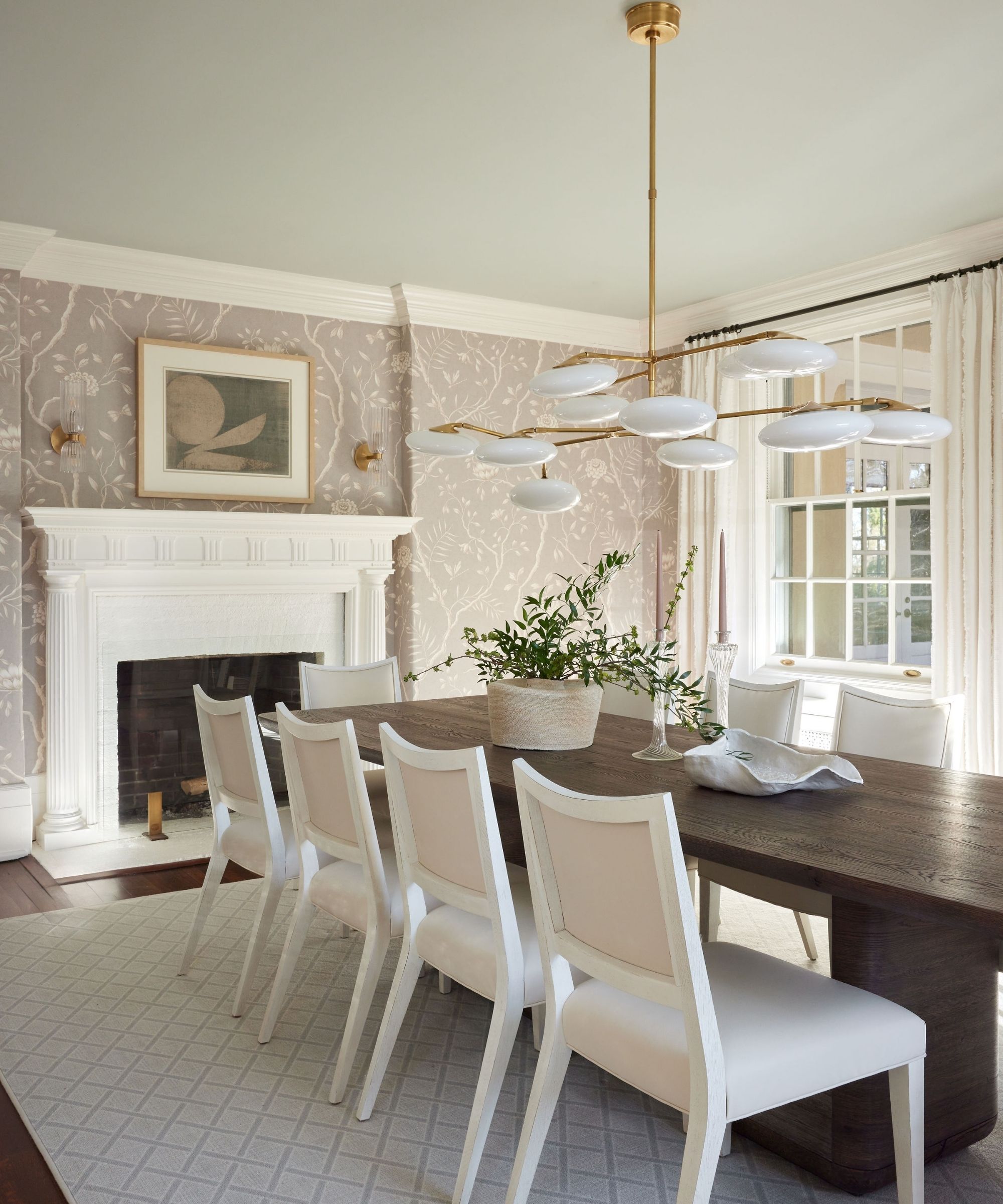Dining room with floral taupe walls, a pale blue ceiling, a long dark wood dining table with white upholstered chairs underneath, a low glass modern pendant light, and a large white fireplace.