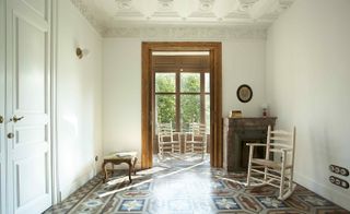 Interior view of a room at Yök Casa + Cultura featuring white walls, white panel doors, a white ceiling with decorative moulding, multicoloured patterned flooring, a fireplace, a coffee table with a book, a sphere wall light, a photo in an oval frame and a off-white coloured rocking chair. There is also a partial view of another room with two chairs and tall windows