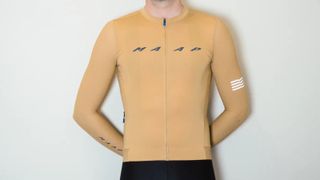 A white man in a tan long sleeve cycling jersey and black cycling shorts stands against a white wall