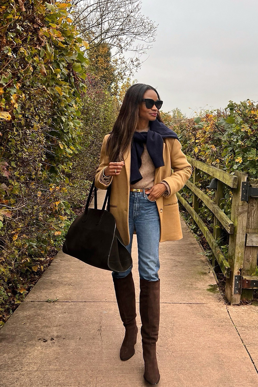 How to wear skinny jeans is shown in a photo of a woman standing outside wearing black sunglasses, a tan blazer, a navy cardigan tied around the shoulders, a camel sweater, a tan belt, a black bowler bag, and skinny jeans tucked into brown suede knee-high boots