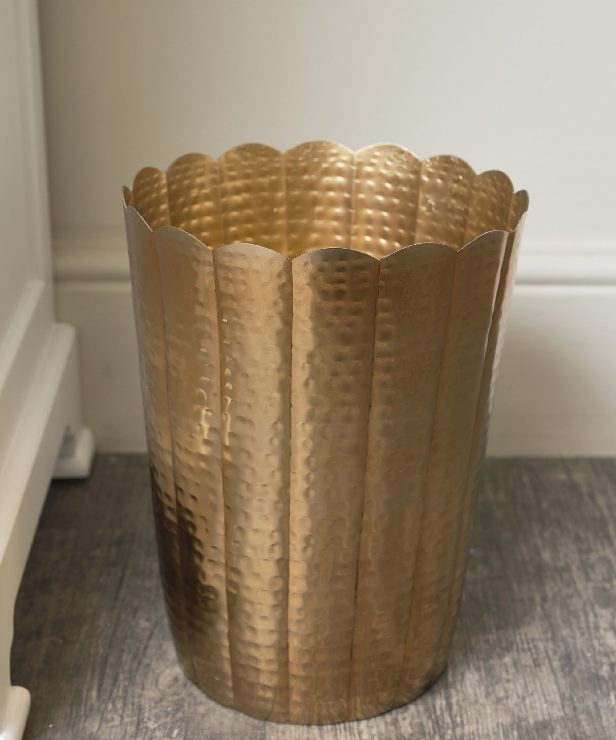 A decorative gold metal bin with a hammered finish and scalloped edge, placed on a wooden floor. Stylish and elegant, it doubles as a waste bin or decorative accent.