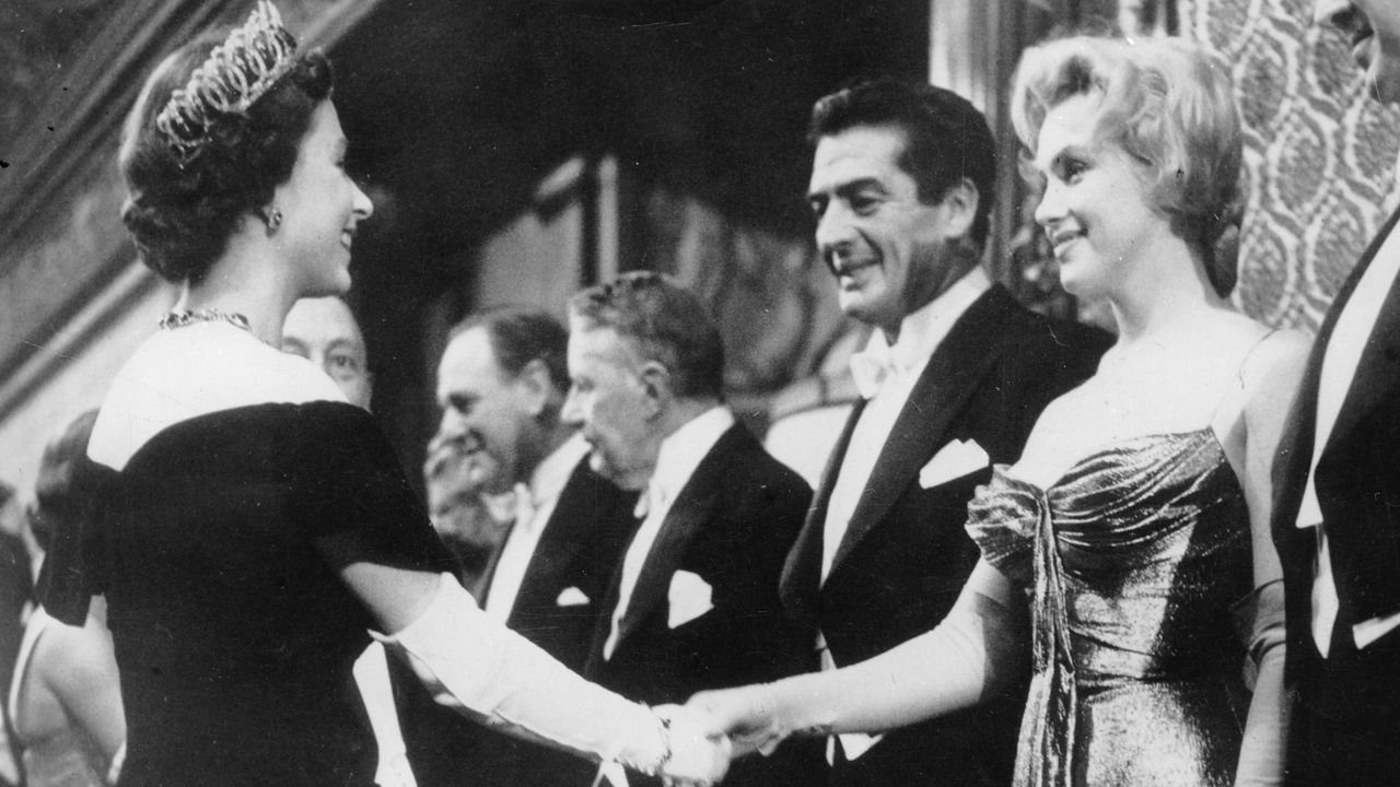 Queen Elizabeth wearing a tiara and evening gown shaking hands with Marilyn Monroe