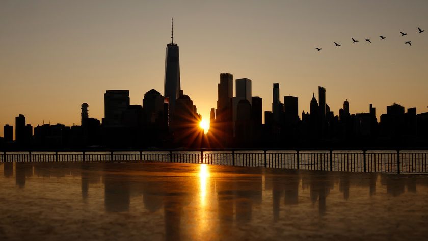 The sun is pictured rising between the skyscrapers of a city skyline. Its light is reflected in a body of water at the bottom of the image, while birds can be seen flying in a &#039;V&#039; formation in the upper left.