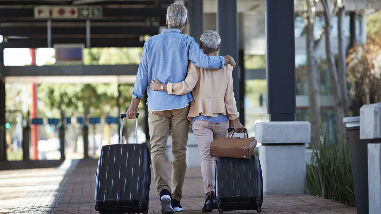 An older couple walk arm in arm outside an apartment with their luggage.