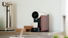 Image of a modern pod coffee machine on a stainless steel countertop. There is a white mug on the machine and boxes of pods next to it.