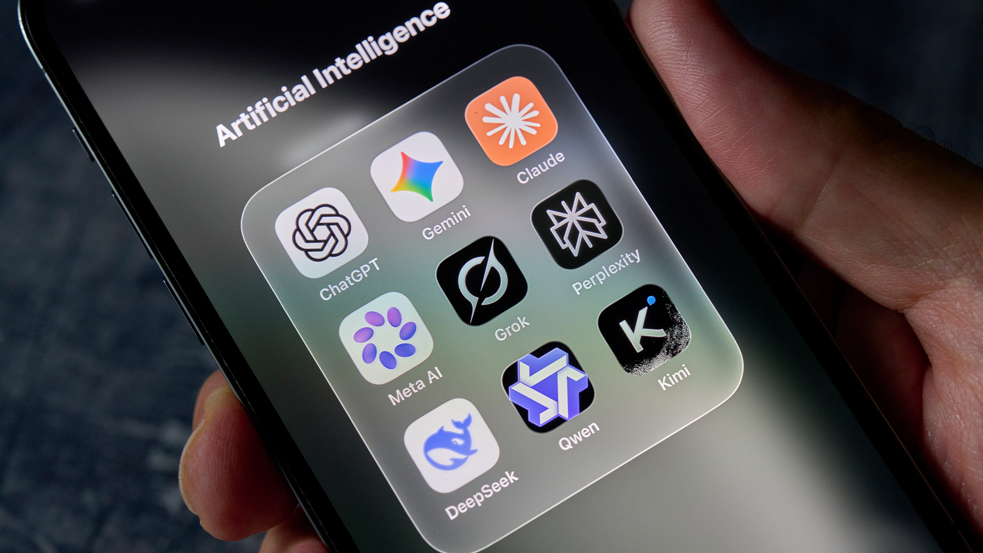 Smartphone screen with selection of AI apps pictured in a dedicated folder titled 'Artificial Intelligence', including ChatGPT, Claude, Grok, Gemini and DeepSeek app symbols.