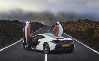 The McLaren 570GT's in white has scissor doors which are both open facing up. The car is in the middle of a road in a mountain valley.