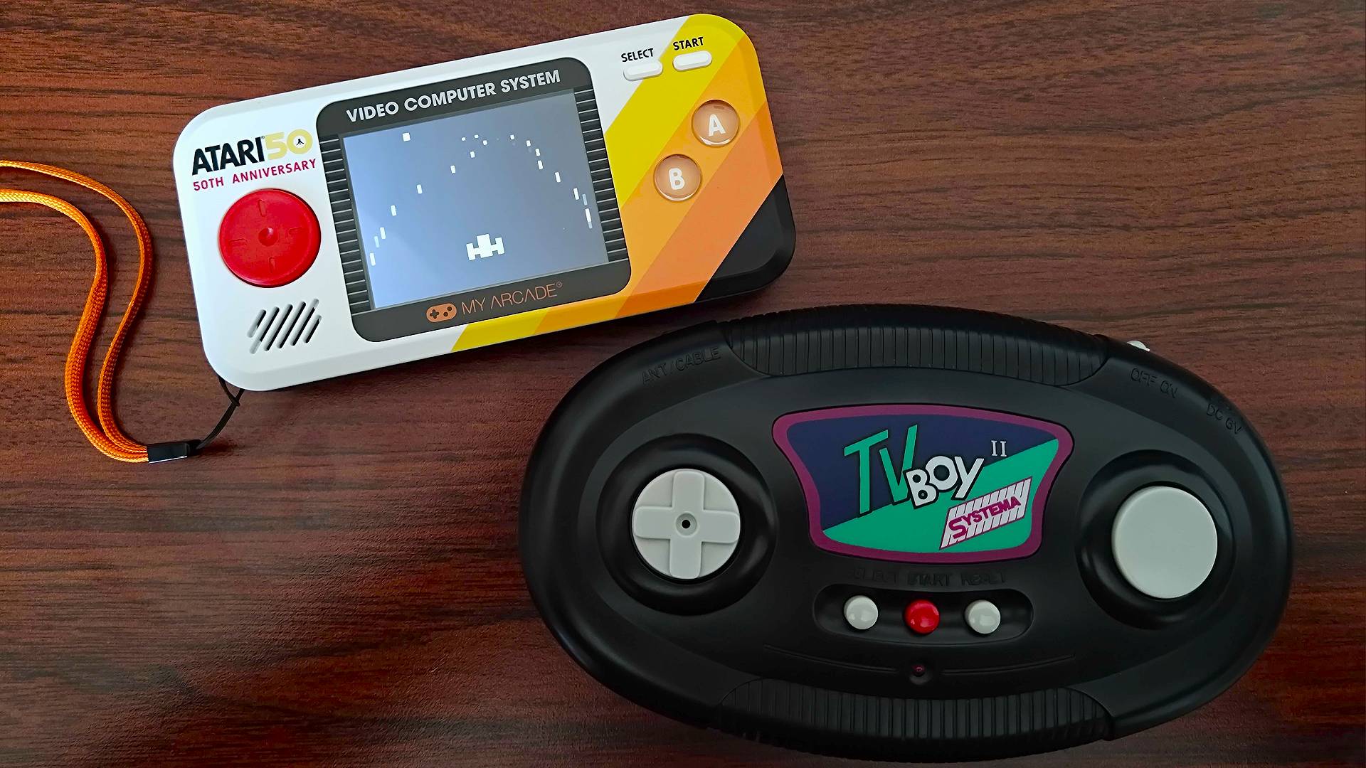 Atari Pocket Player Pro handheld on top left and Systema TV Boy on bottom right with both sitting on woodgrain desk.