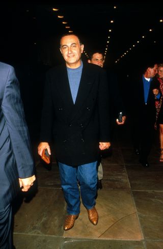 Dodi Fayed walking towards the camera in a black blazer, blue shirt, jeans, and boots