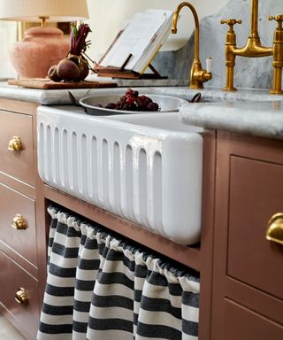 close-up of a white ceramic farmhouse sink with a ribbed front apron, set against a marble countertop. The sink is framed by brown cabinetry with gold hardware and a striped fabric curtain. A brass tap, cutting board with vegetables, and an open cookbook are visible.