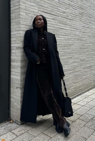 how to wear velvet is shown in a photo of a woman standing outside wearing a long black coat over a dark brown velvet jacket, dark brown velvet trousers, a black Prada bag, silver jewelry, and black tabi-toe ankle boots