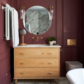 A purple-painted bathroom with a round wall mirror above a wooden bathroom cabinet