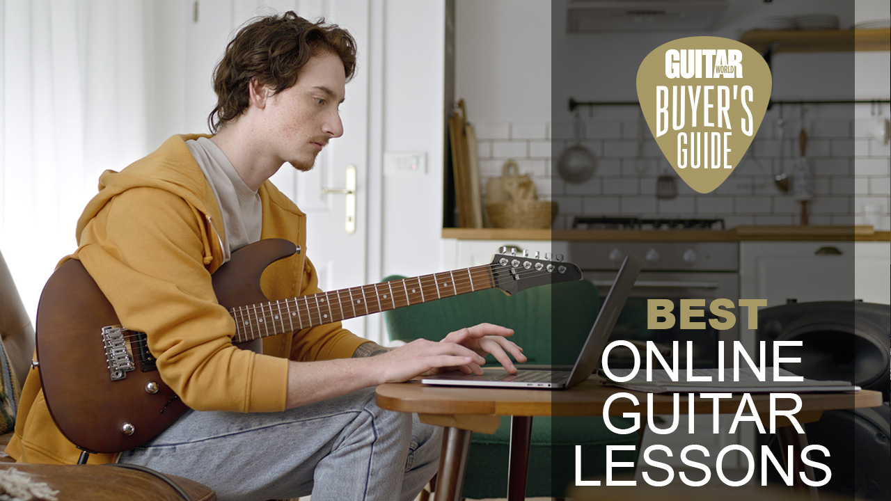 Man in yellow jumper holding a natural finish electric guitar types into a laptop