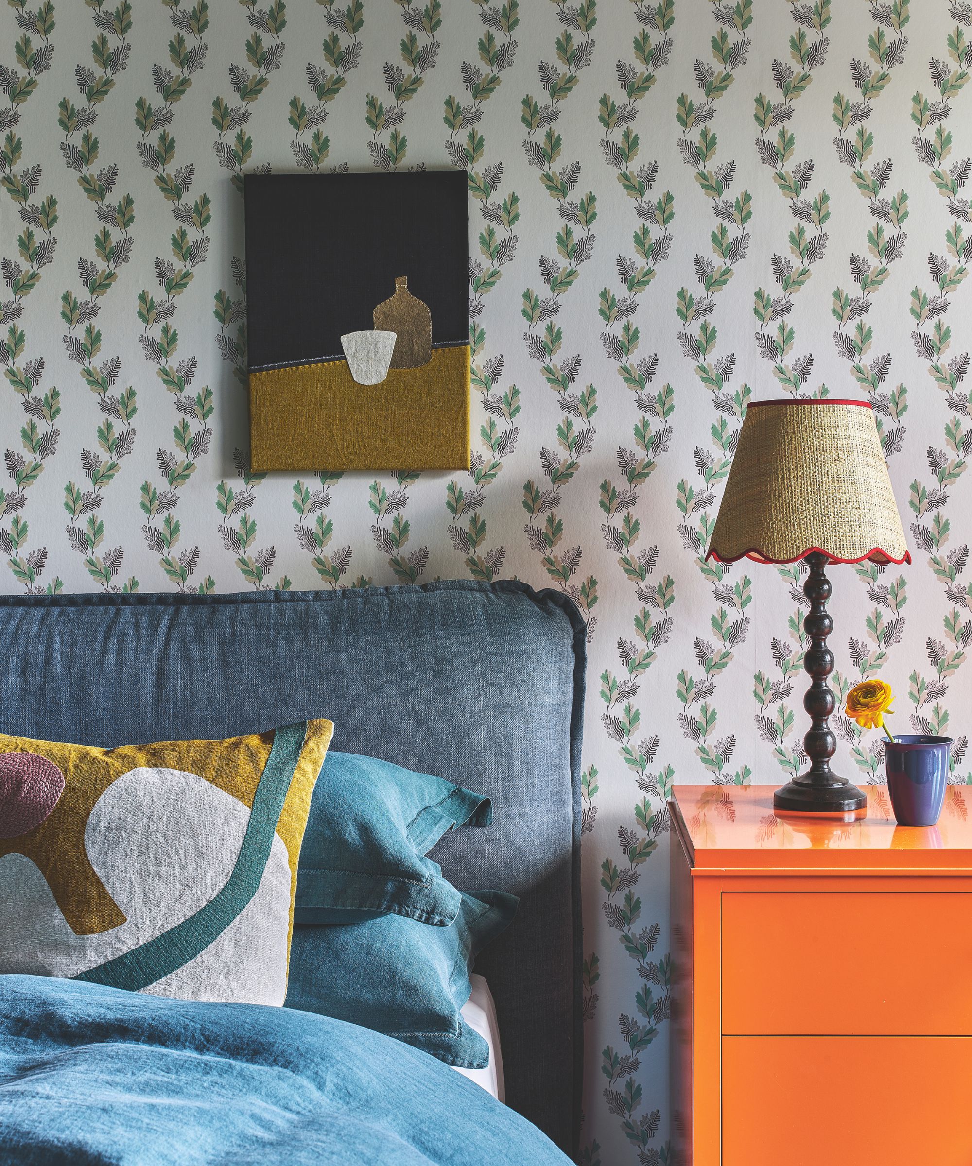 A floral bedroom with blue upholstered bed with blue bedding and patterned cushion, a bright orange chest of drawers with a brown lamp with rattan scalloped shade, and a blue vase with yellow flower.