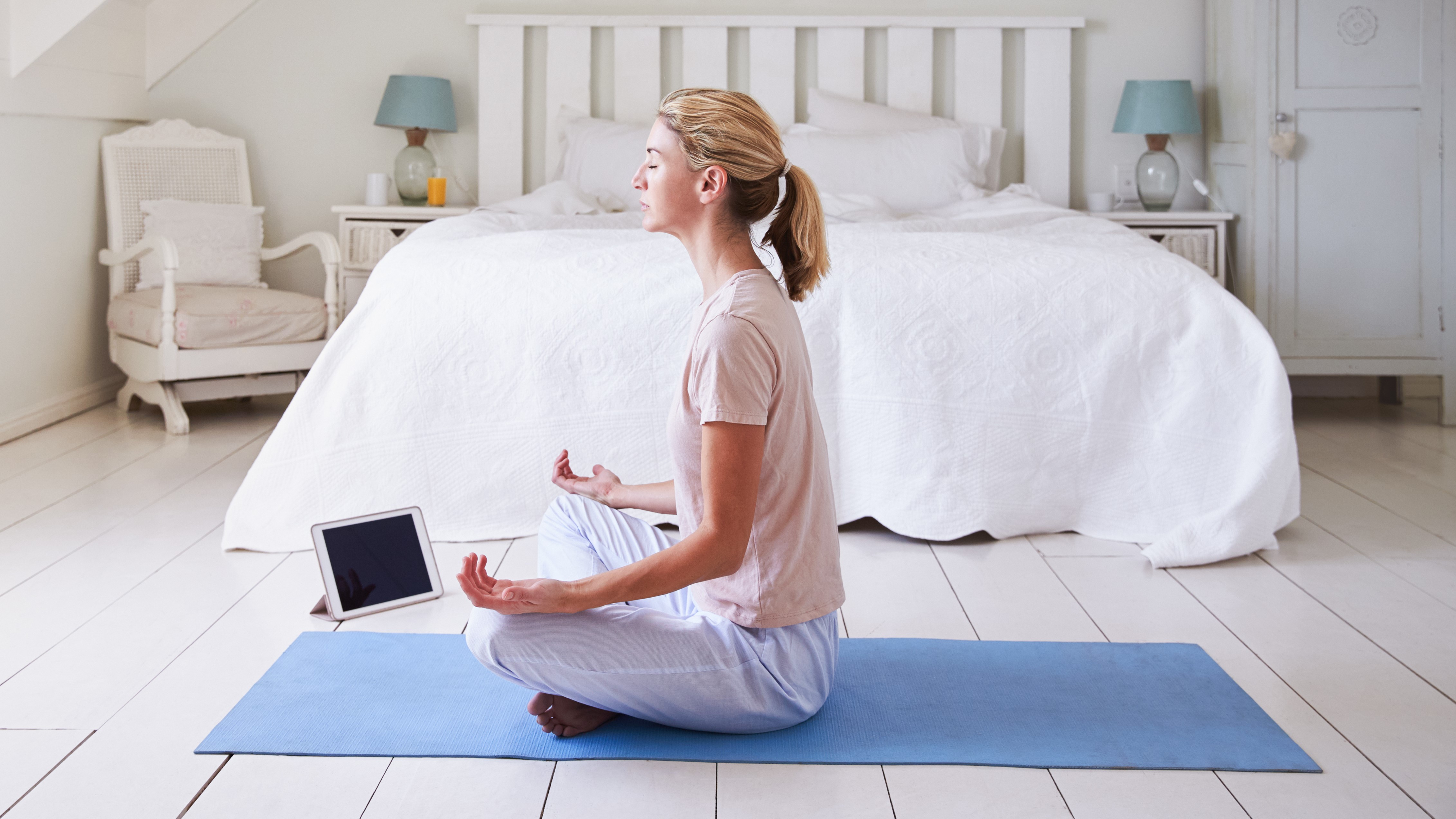 Женщина использует приложение для медитации, сидя на синем коврике для йоги перед своей белой кроватью.