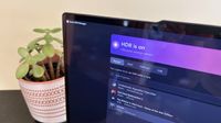 A monitor displays a purple Smart HDR Manager settings menu with HDR enabled and a list of games, beside a potted plant in a bright room.