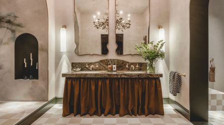 A bathroom interior with checkered beige floors, a large, two-basin sink with a brown and green marble top and a curtained shelving base, two antique mirrors placed atop it, and arched alcoves.