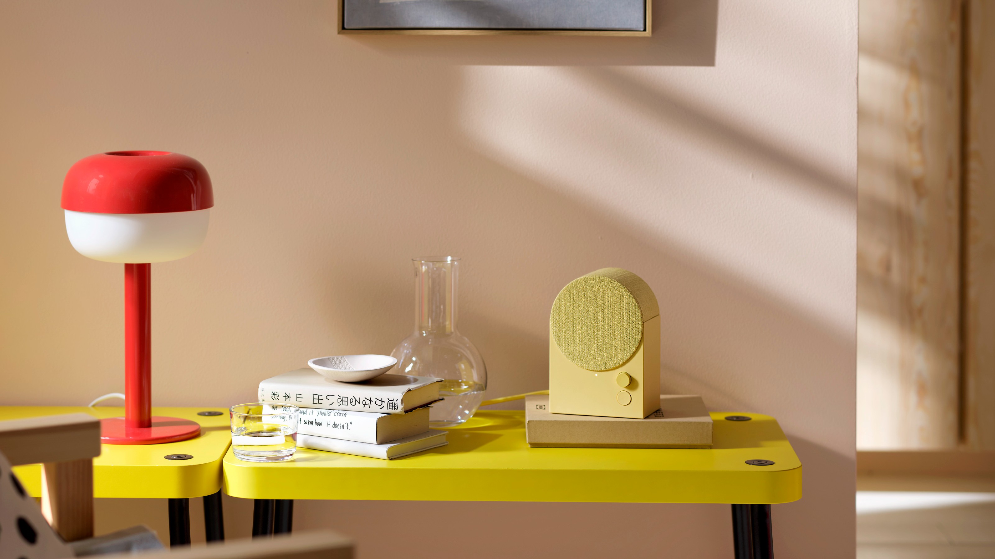 The IKEA NATTBAD bluetooth speaker in yellow on a yellow side table against a pink wall and next to a red IKEA table lamp