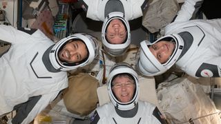 four astronauts in white and gray spacesuits floating in four different directions with their heads pointed to center