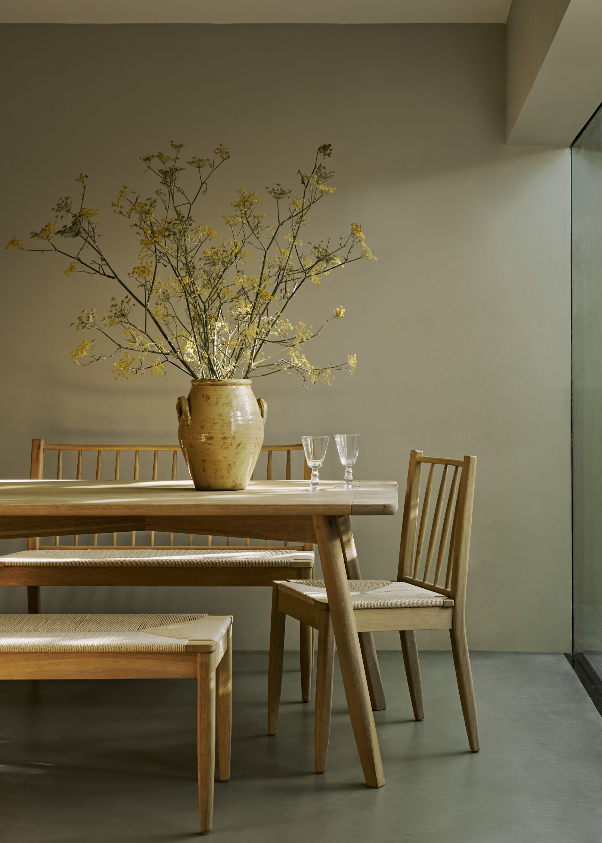 Corner of a modern dining room with earthy beige walls, a wooden dining table with a vase of foliage, and a woven bench.