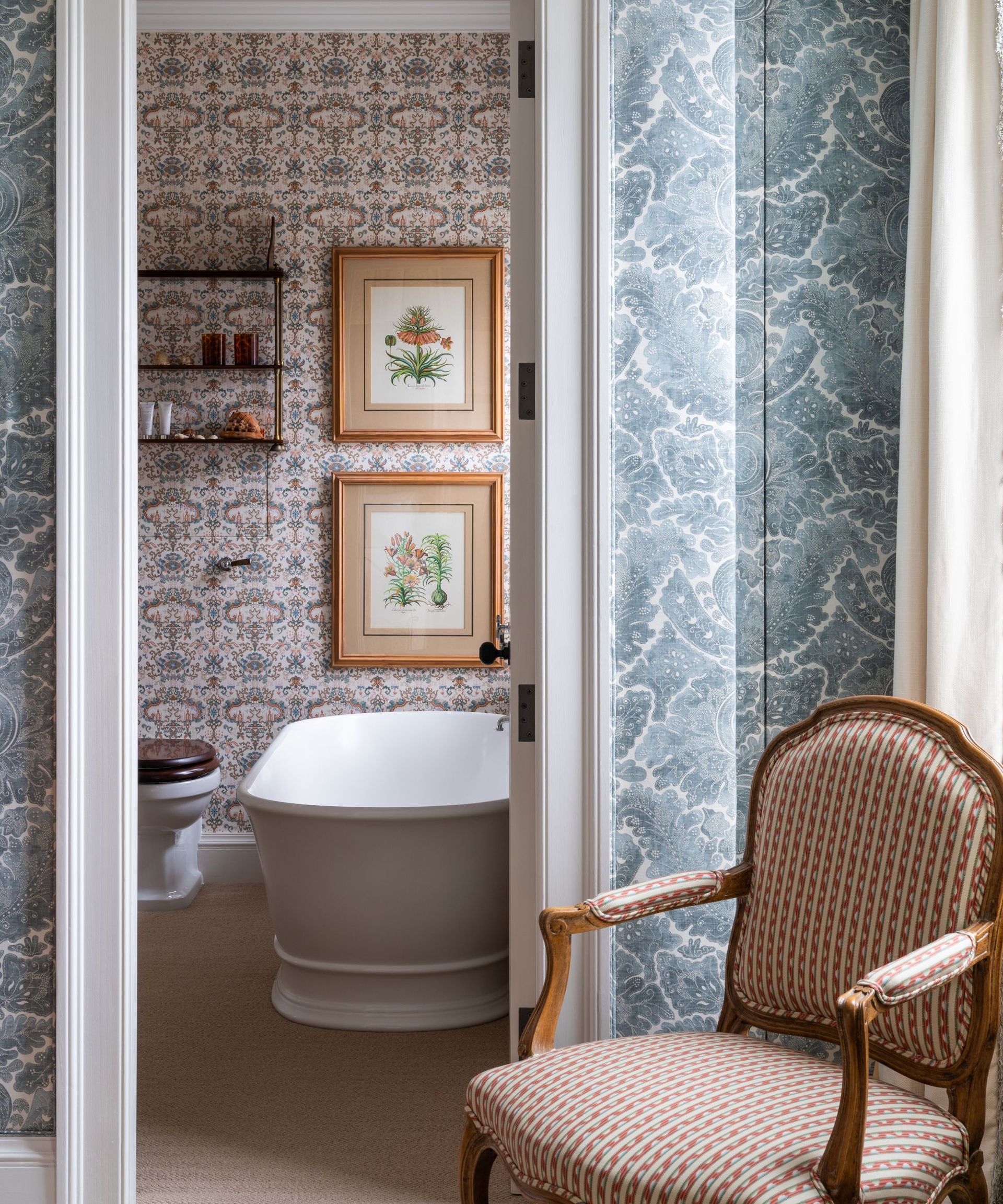 Wallpapered bathroom with vintage flower prints, bathtub and shelf and vintage upholstered chair