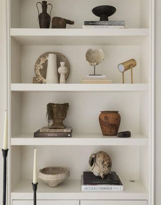 Close-up image of a white open shelving unit that is decorated with different decor items. There are small, ceramic vases, stacks of books, candle sticks, and art pieces.