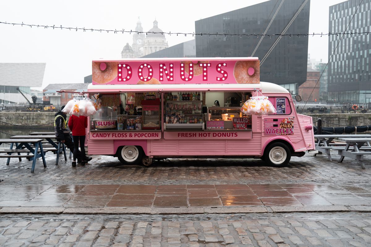 A pint donut truck in on a grey rainy day with large buildings in the background
