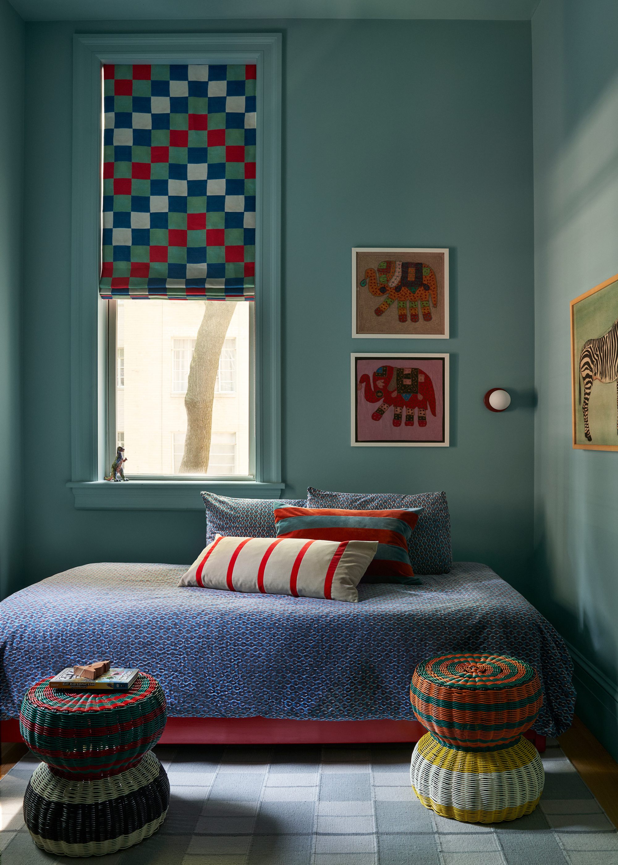 A children's bedroom with framed elephant art, a colorful low bedscape, checkered blinds, woven stools, and a wall sconce with a red base