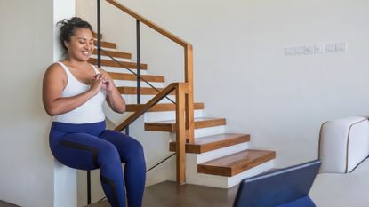 A woman performs the wall sit exercise at home. She presses her back against a wall with her upper legs horizontal and her knees bent to 90°. She wears a white tank top, blue sports leggings, and is smiling 