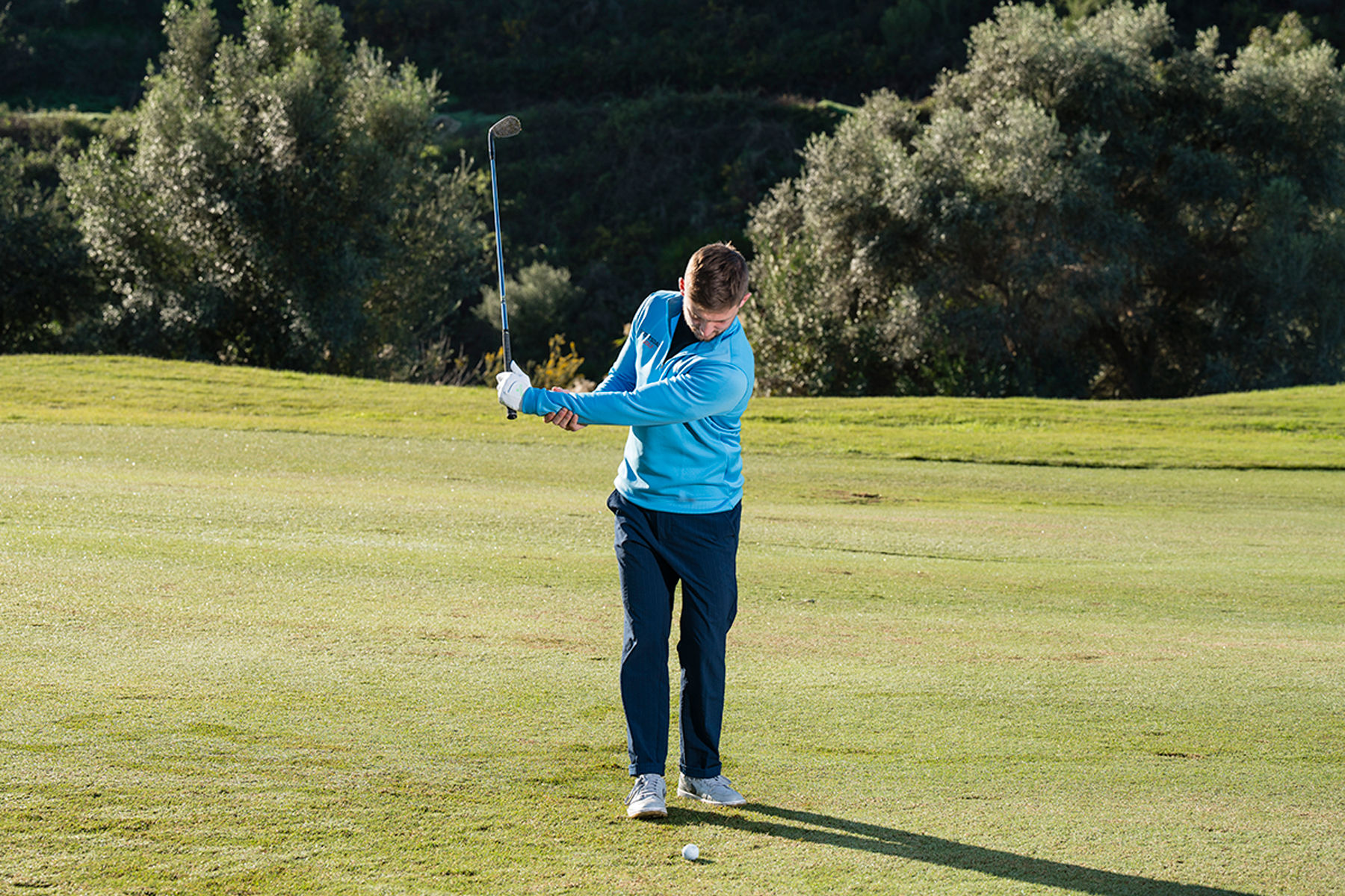 PGA Pro Alex Elliott demonstrating the gravity drill, with a wedge in his glove hand and his lead arm outstretched parallel to the ground in the backswing, supported by his right arm to prevent gravity pulling the club down to the ground