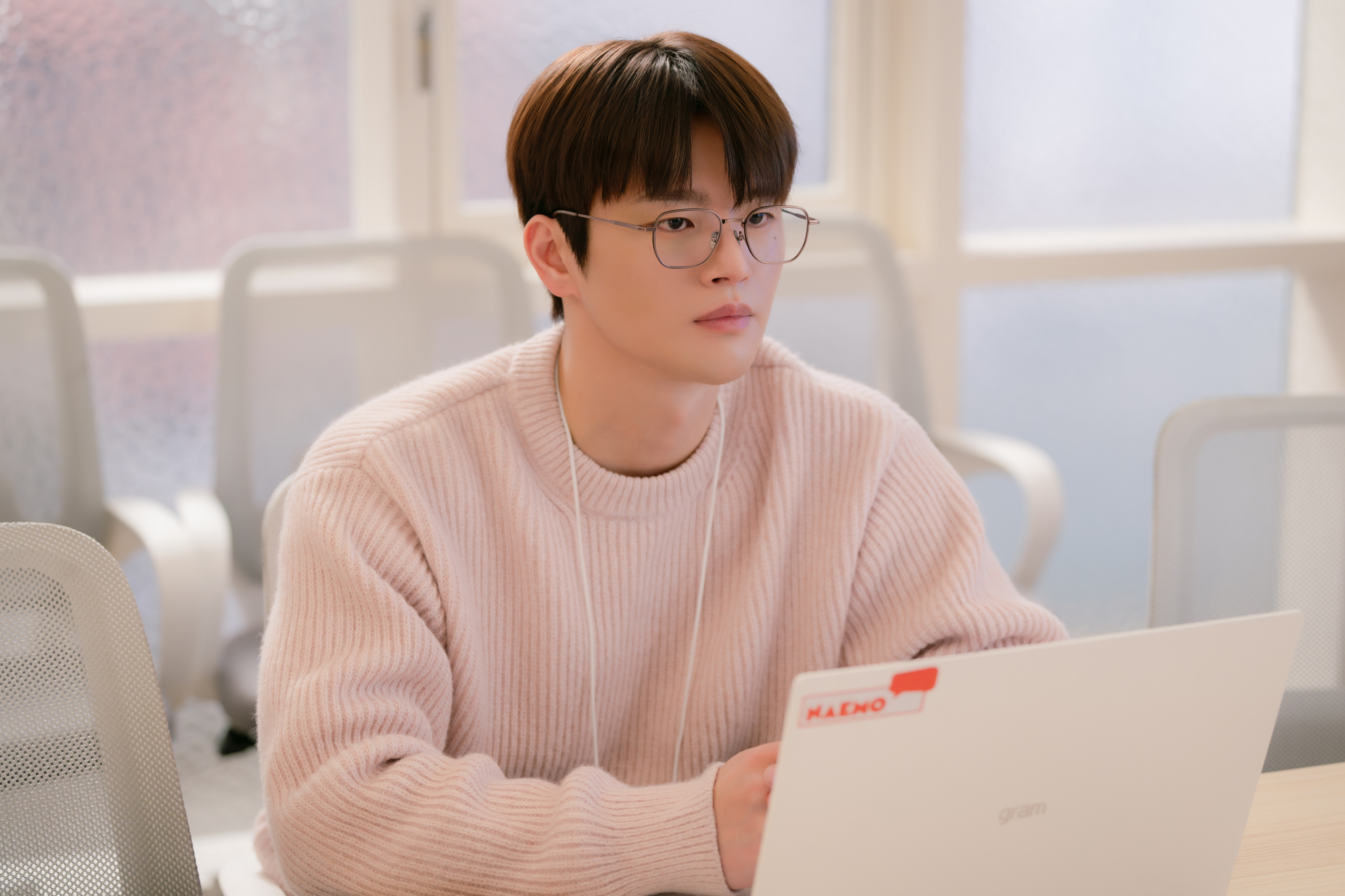 A man (Seo In-guk as Park Gyeong-nam) wears glasses, a pink sweater, and a white lanyard as he sits in front of a laptop in a conference room, in the Netflix K-drama 'Boyfriend on Demand.'
