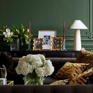 A green living room with a brown sofa that has animal print throw pillows on it. There is a vase of white hydrangeas by the sofa and a black console table behind the sofa. There is a white lamp, gold picture frames, and red candles on the black console.