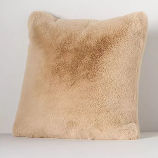 A furry beige colored square pillow against a white backdrop