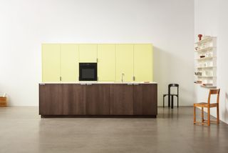 A large kitchen display with tall canary yellow cupboards and a wooden island set against a white wall. A whilte floating shelf on the right next to a black shair and a wooden chair.