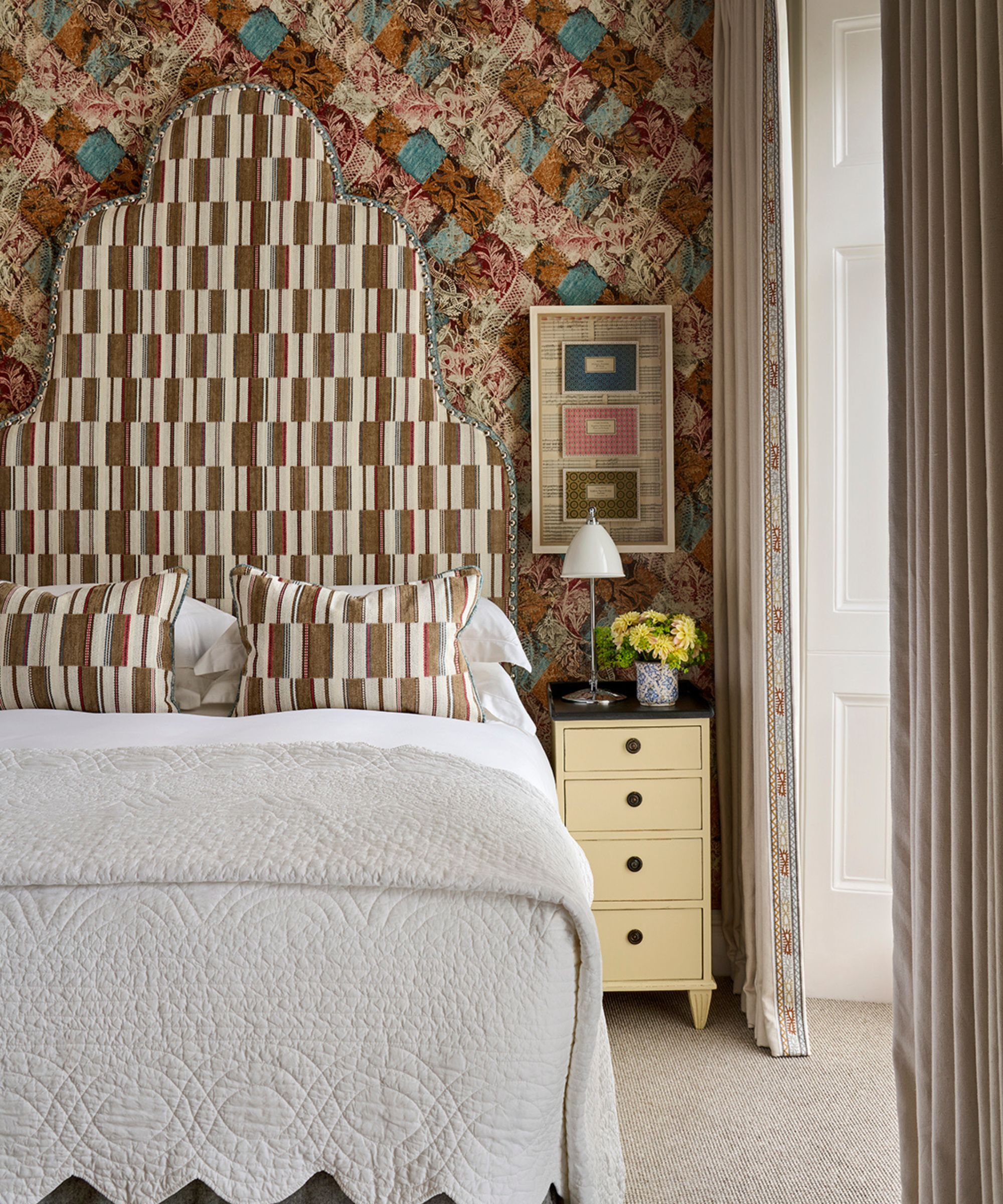 Bedroom with brown chequered wallpaper, a tall brown geometric headboard and matching throw pillows, white bedding, a yellow side table with a lamp, and a large window with long drapes.