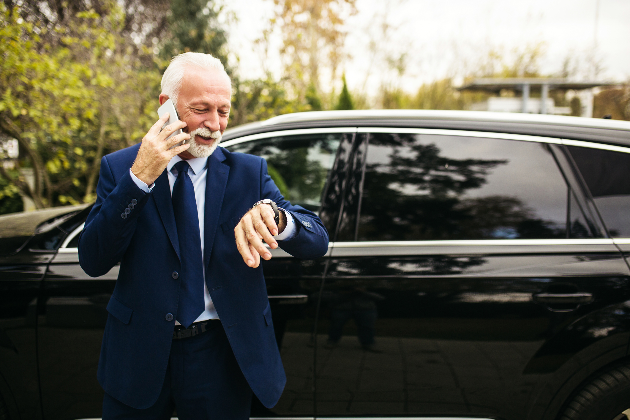 Senior adult driver standing and talking on the phone next to luxury car