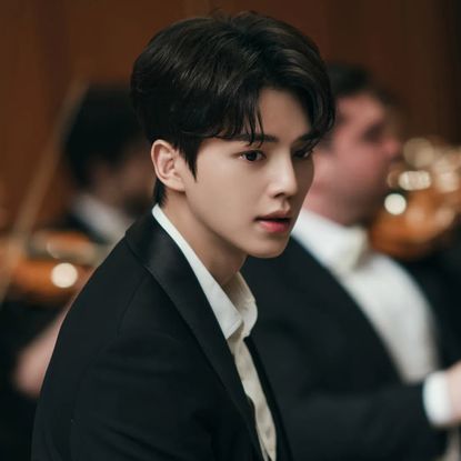 A Korean man (actor Song Kang) wears a suit while sitting in front of a blurred orchestra, for the K-drama 'Four Hands, Two Sonatas.'