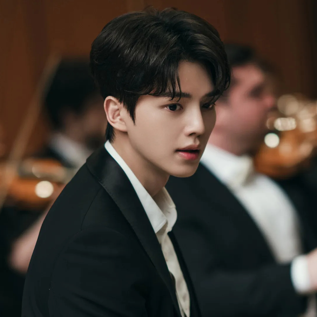 A Korean man (actor Song Kang) wears a suit while sitting in front of a blurred orchestra, for the K-drama 'Four Hands, Two Sonatas.'