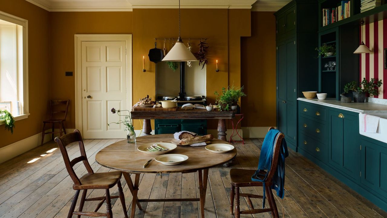 Mustard yellow and dark blue kitchen diner with hard floors and striped red and white backsplash. There are units on the right, and a window to the left, with a table in the background alongside a range cooker inset in an alcove
