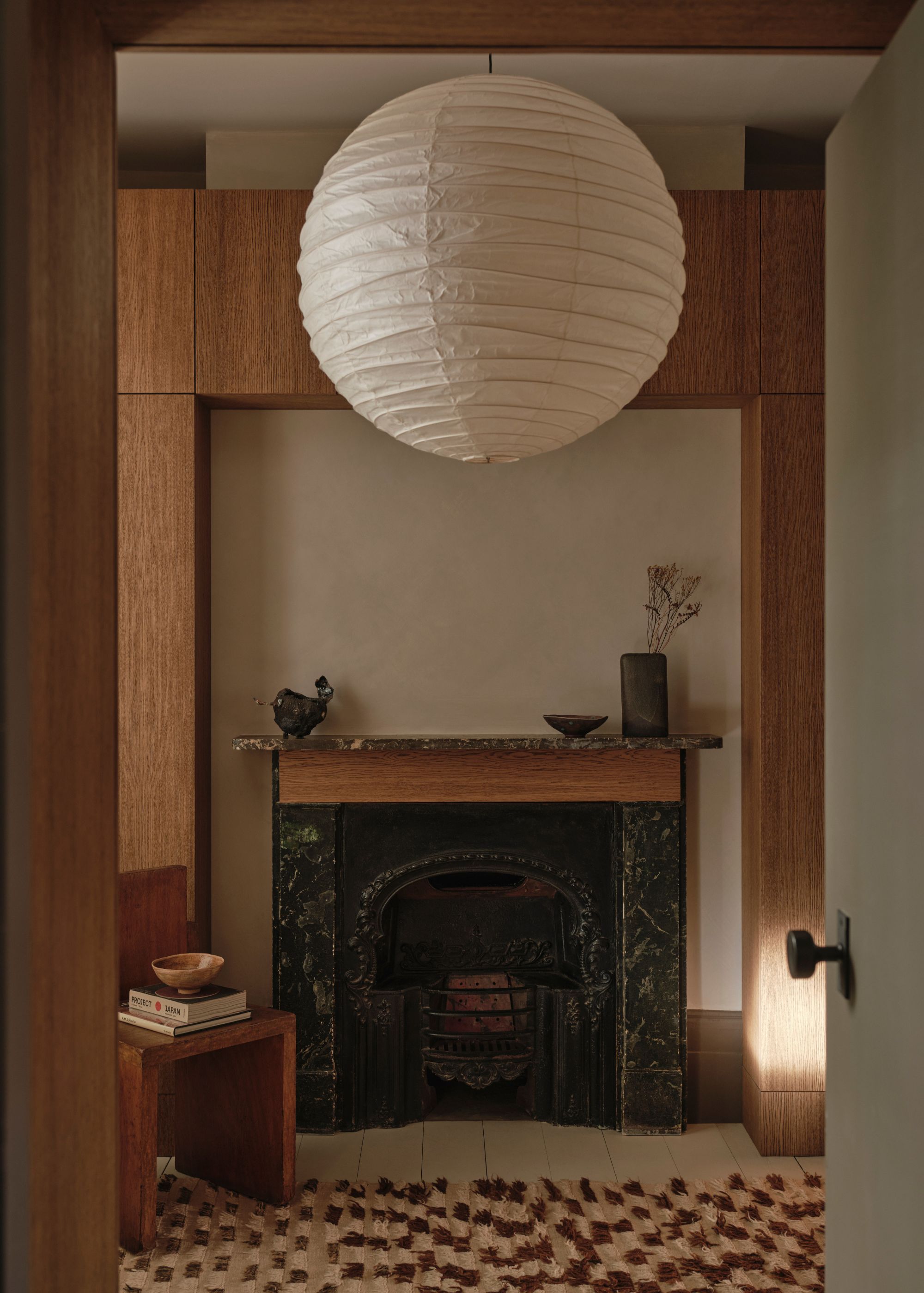 A living room with a large paper lamp pendant, a stone fireplace, a vase of stems, ceramic bowls, a patterned rug, coffee table books, and a wood chair