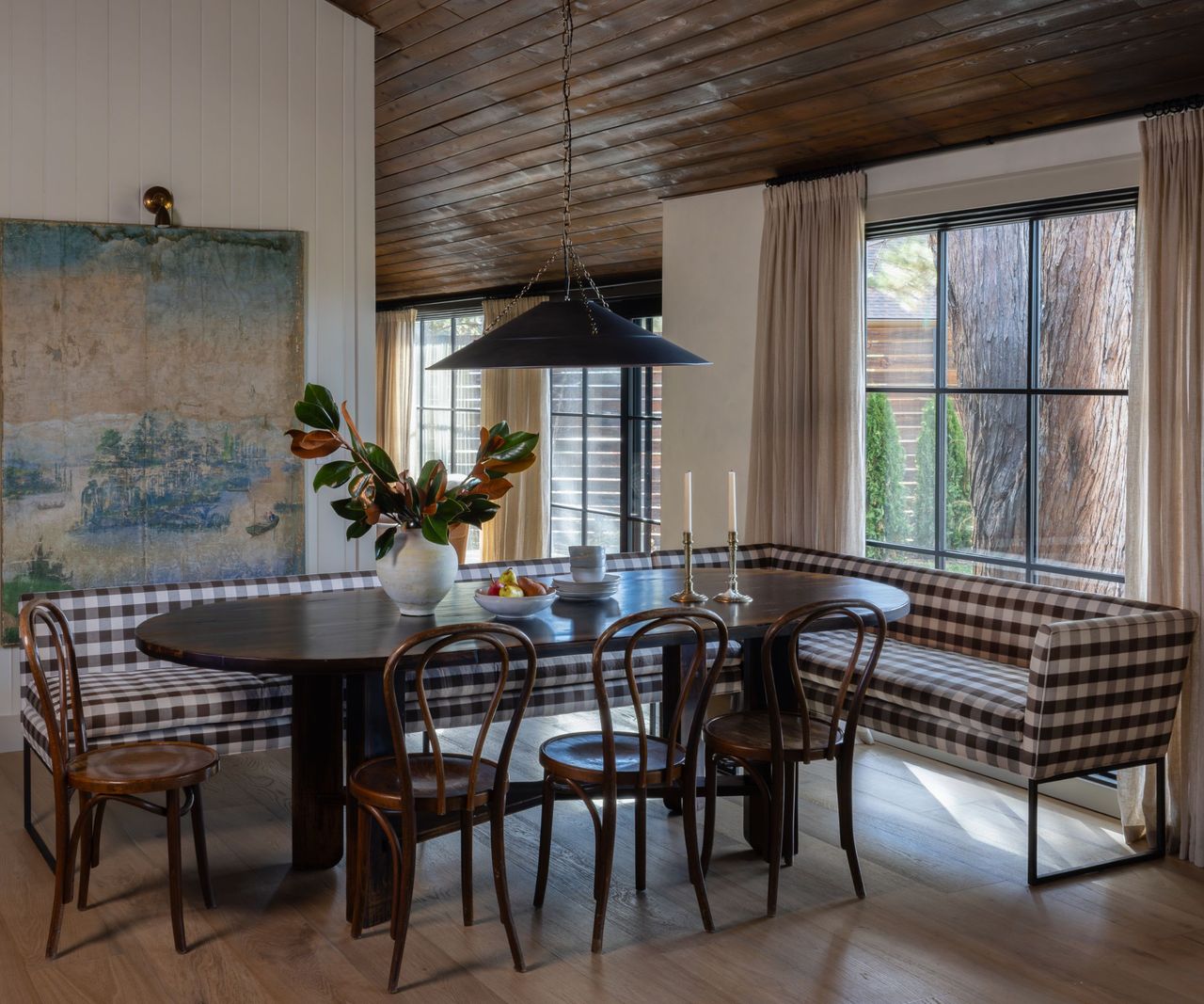 A dining nook with a dark wooden oval table surrounded by wooden chairs and a checkered banquette. A large vintage wall hanging is on one wall and a black pendant light hangs above the table