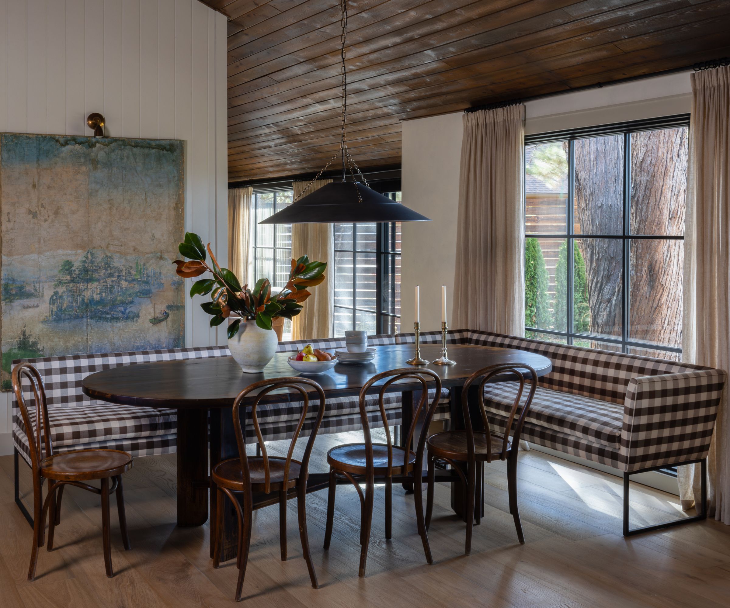A dining nook with a dark wooden oval table surrounded by wooden chairs and a checkered banquette. A large vintage wall hanging is on one wall and a black pendant light hangs above the table