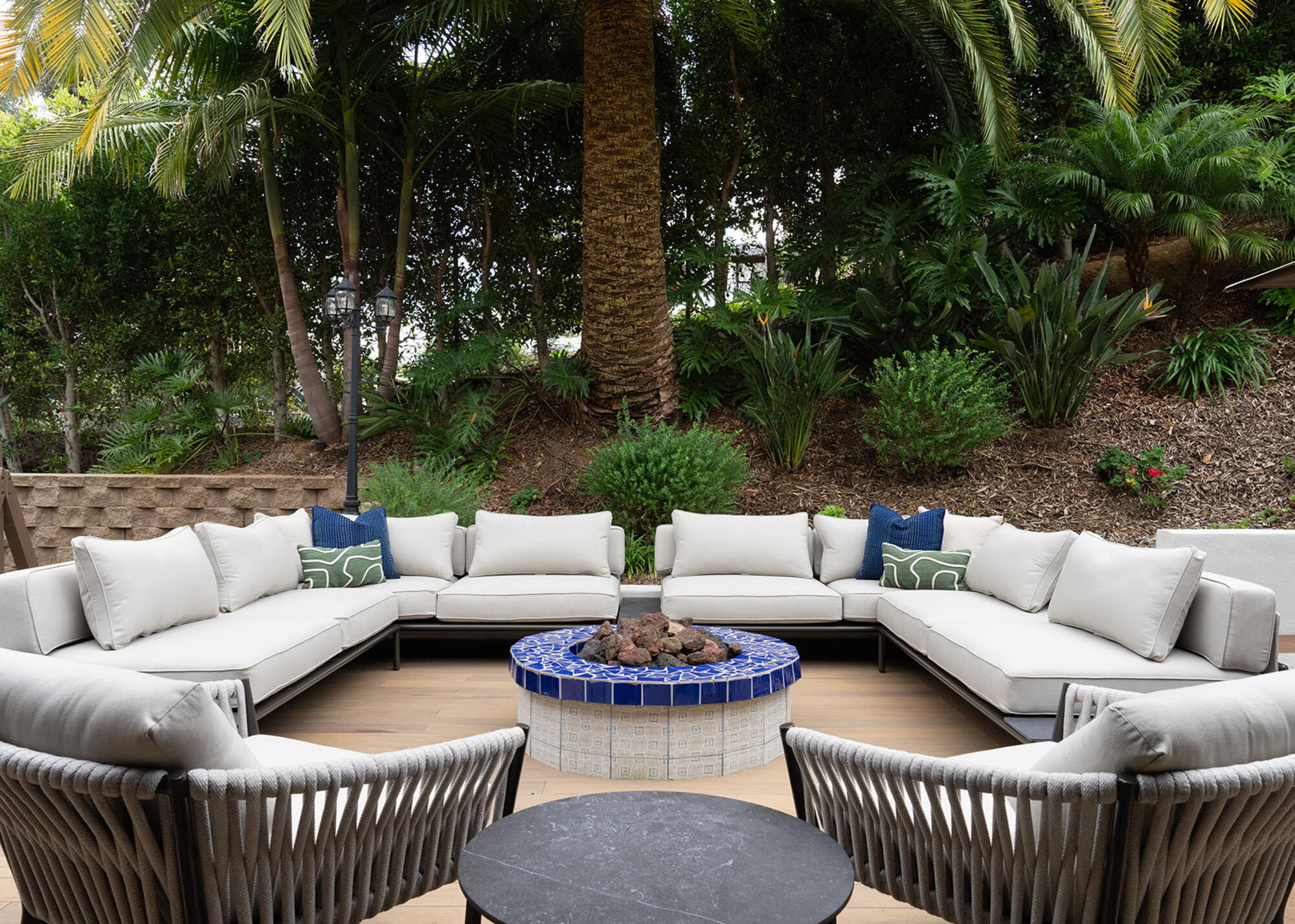 An outdoor conversation pit with a large sofa and garden chairs surrounding a blue tiled fire pit