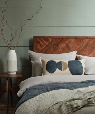 bedroom with pale green panelled wall, bed with darker wood headboard, cushion with dark grey and green circle pattern