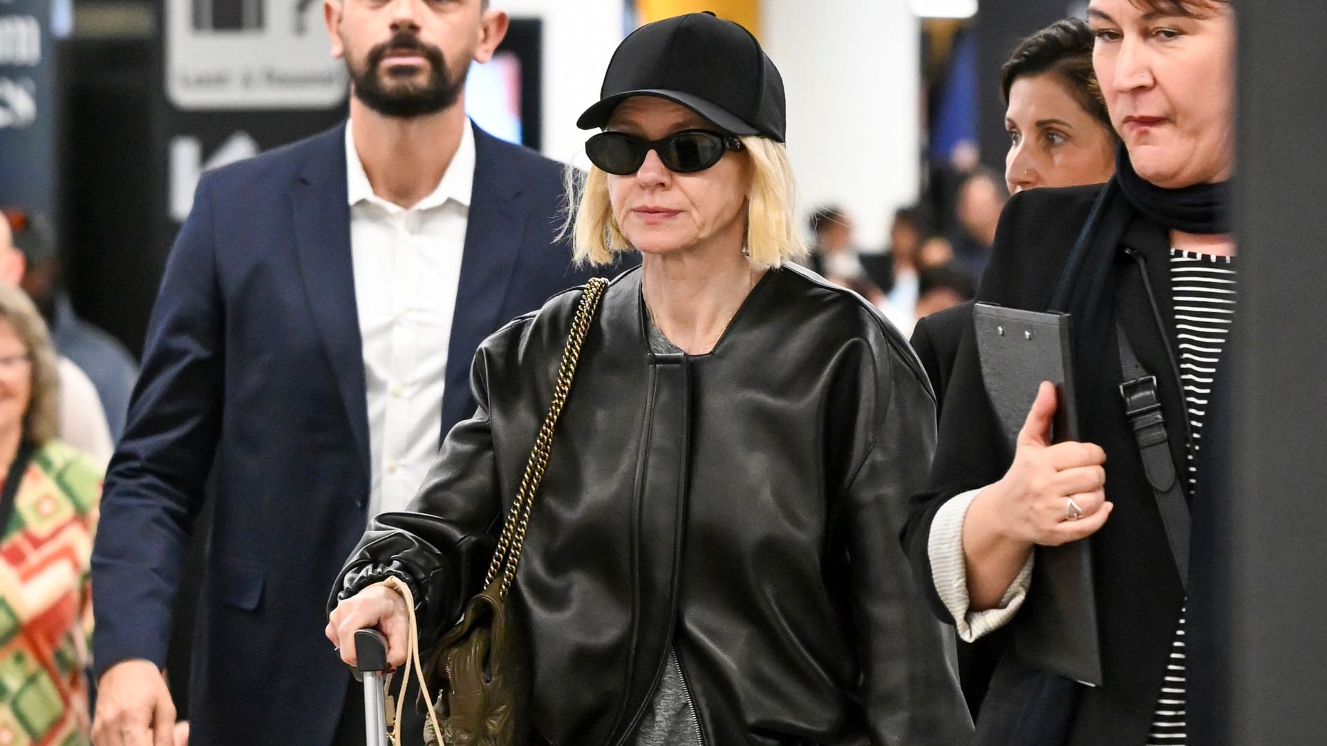 Naomi Watts walks through the airport wearing grey track pants with a black leather bomber jacket, red trainers, a black cap and black sunglasses.